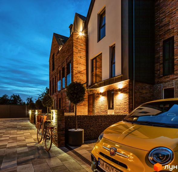 Котеджне містечко КМ Wunder Haus з цегляним фасадом та підсвіткою. Поруч розташовані велосипед та жовтий автомобіль на бруківці.