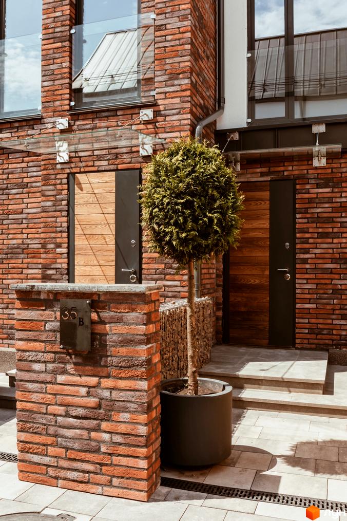 Фасад котеджного містечка КМ Wunder Haus з цегляною кладкою та дерев'яними дверима. Поруч розташована прибудинкова територія з кущом та бруківкою.