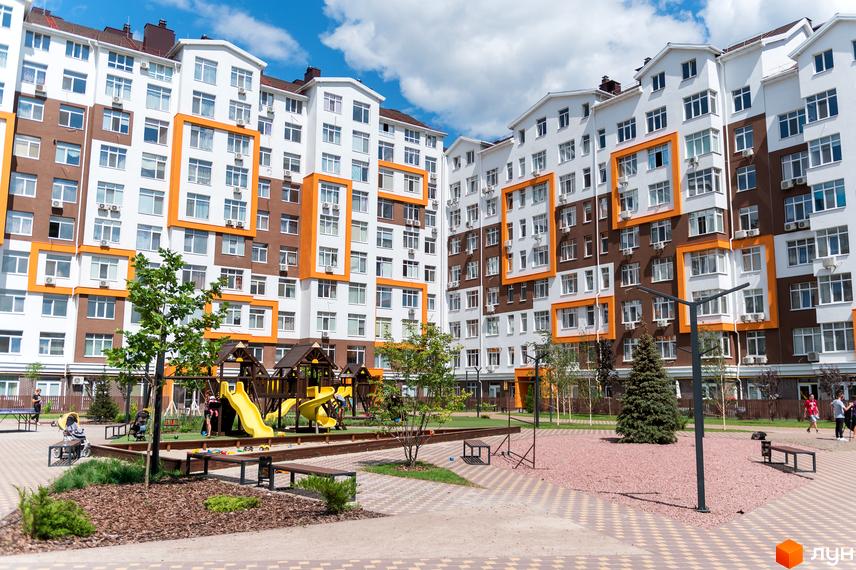 Житловий комплекс Orange Park з білими та коричневими фасадами, прикрашеними помаранчевими елементами. На прибудинковій території розташований дитячий майданчик та зелені насадження.