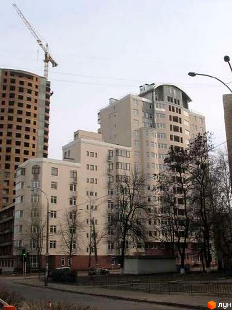 Світлий житловий комплекс з округлими балконами та вигнутим дахом, поруч недобудований цегляний будинок з краном. На передньому плані вулиця з деревами без листя та ліхтарем.