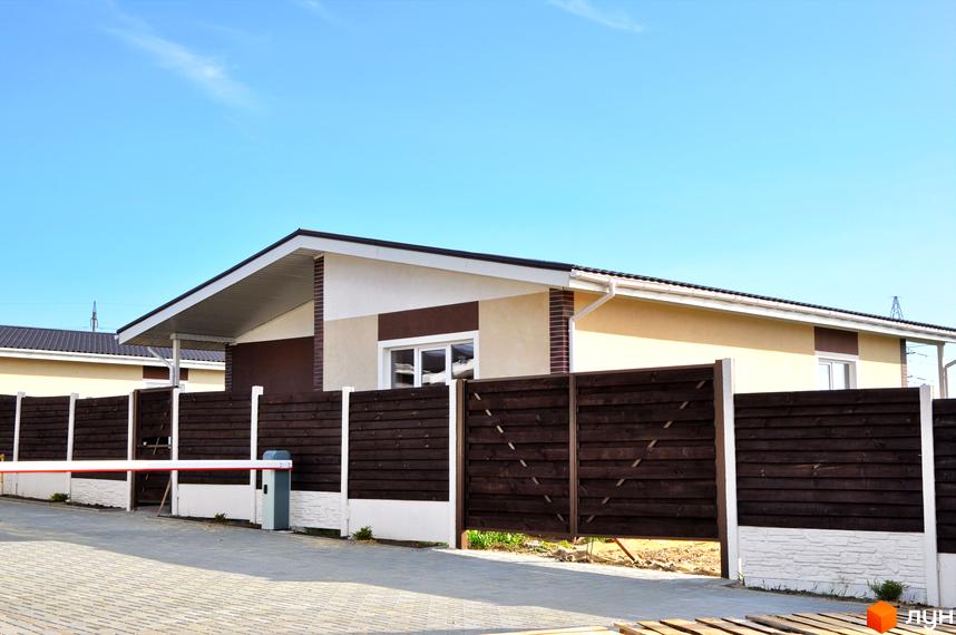 Котеджне містечко КМ Данилівський: одноповерхові будинки з коричневим та бежевим фасадом, білою покрівлею та темним дерев'яним парканом. На території встановлено шлагбаум.