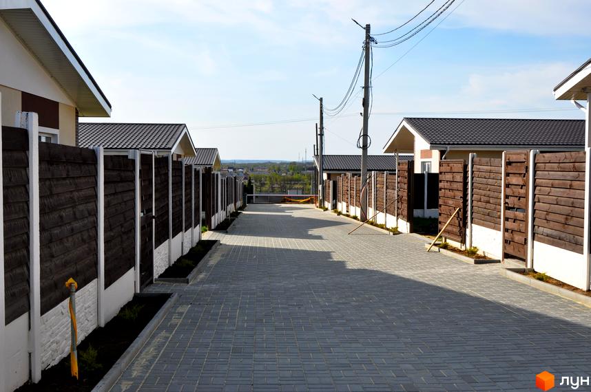 Котеджне містечко Данилівський: вулиця з бруківкою та парканами, що веде до будинків з темними дахами. Прибудинкова територія з доглянутими газонами та вуличним освітленням.