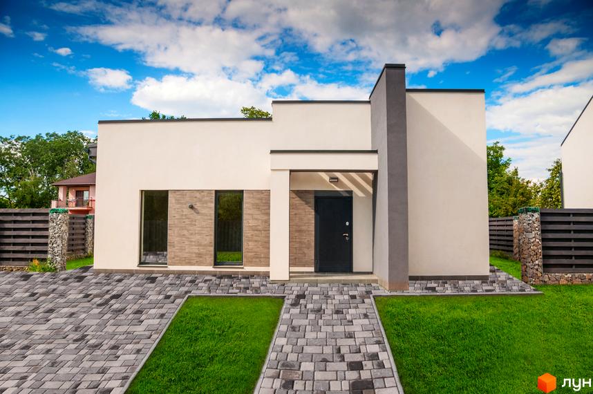 Одноповерховий будинок з білими стінами та темними вікнами. До будинку веде доріжка з бруківки, навколо зелена трава.