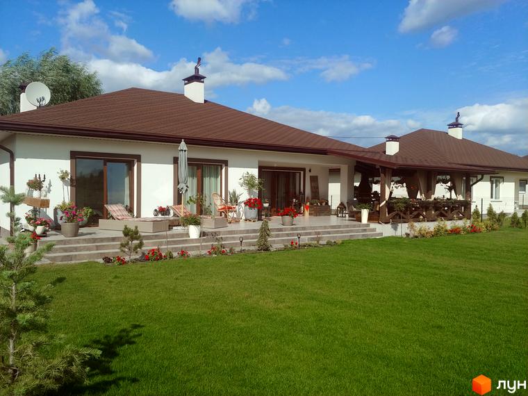 Котеджне містечко Маєток: одноповерховий будинок з коричневим дахом, терасою та зеленою галявиною. Навколо будинку висаджені квіти та дерева.