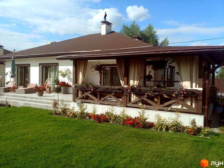 Котеджне містечко КМ Маєток: білий двоповерховий будинок з коричневим дахом, терасою та квітами на прибудинковій території. Зелений газон та квіти перед будинком.