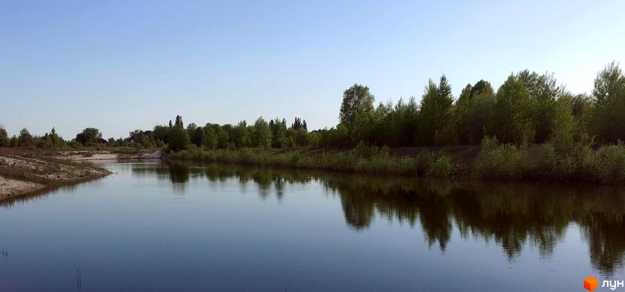 КМ Маєток: мальовничий краєвид з річкою та зеленою зоною. Комплекс розташований біля води, оточений деревами та кущами.