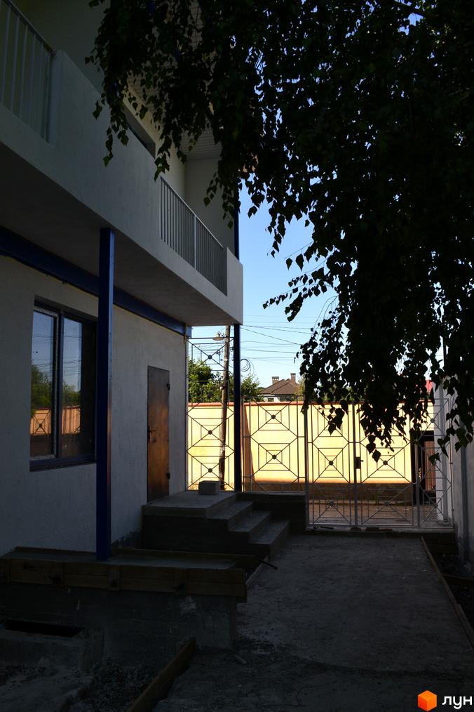 Зображення житлового будинку з білими стінами та балконом. Перед будинком розташована прибудинкова територія з воротами та деревами.