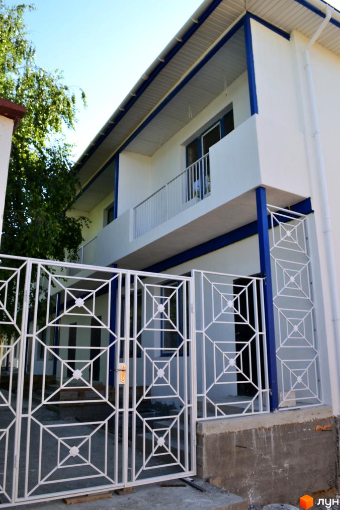 Зовнішній вигляд будинку в ЖК Романтичний з білими стінами, синіми акцентами та кованим парканом. Видно балкон та зелені насадження.