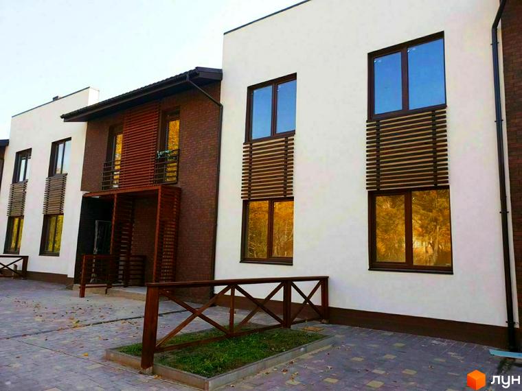 Фасад новобудови з цегляним оздобленням та великими вікнами, що відображають дерева. Прибудинкова територія з дерев'яною огорожею та бруківкою.