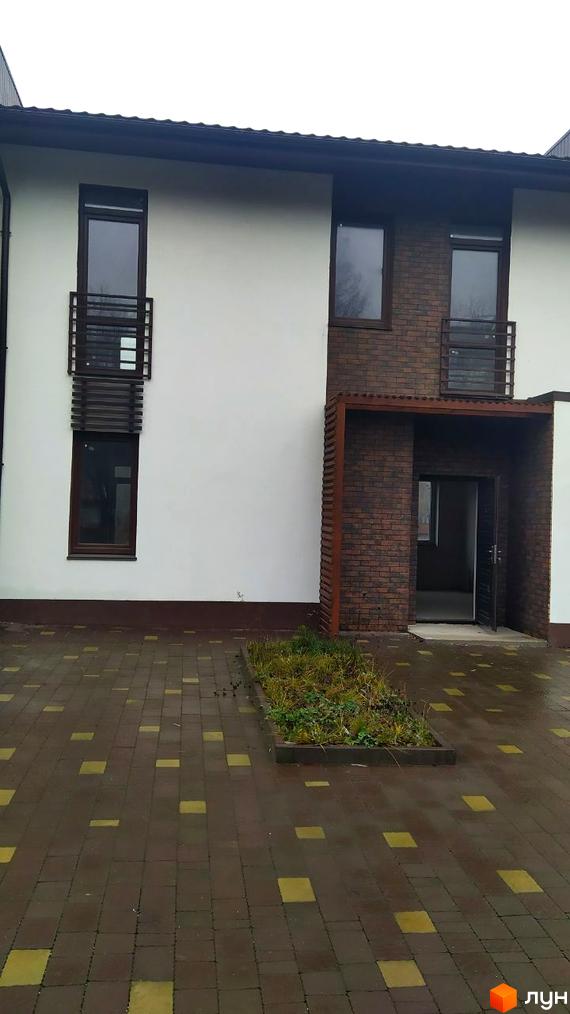 Будинок з білими стінами та коричневою цегляною вставкою, вікнами та балконами. Прибудинкова територія вимощена плиткою з зеленою ділянкою.