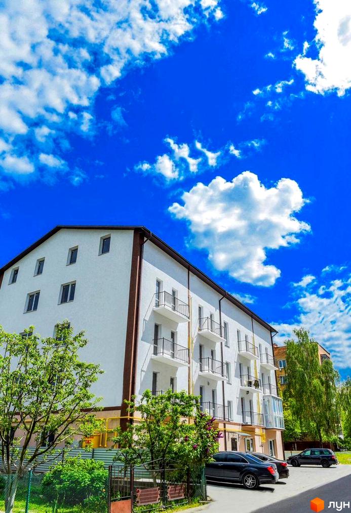 Зовнішній вигляд білої новобудови з балконами та зеленими деревами на прибудинковій території. Простір перед будинком з автомобілями та огорожею.