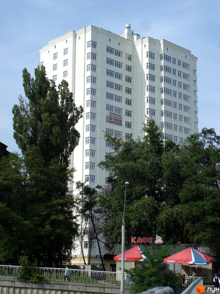 Житловий комплекс Затишний дім – біла багатоповерхівка з численними вікнами на тлі блакитного неба. Навколо будівлі зелені дерева та елементи міської інфраструктури.