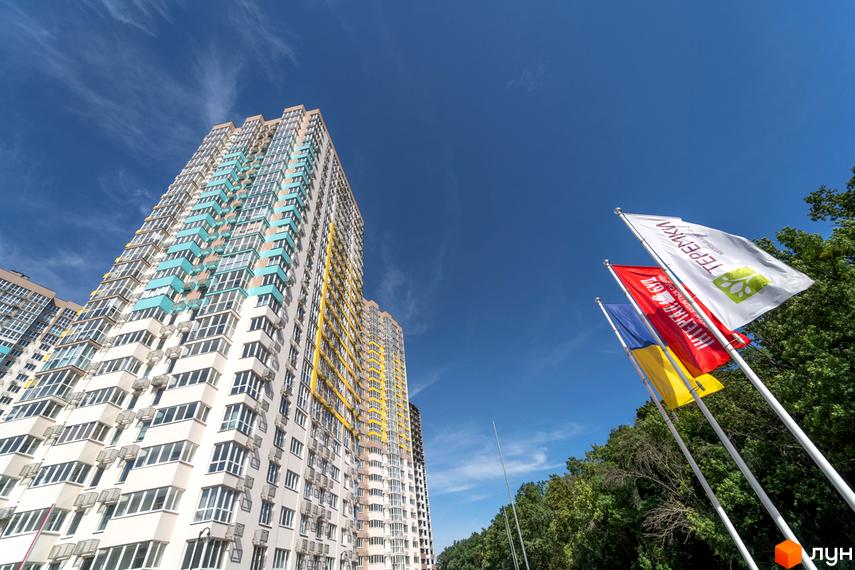 Житловий комплекс Теремки. Багатоповерхові будинки з білими та блакитними фасадами на фоні ясного неба. Поруч майорять український прапор та білий прапор з логотипом проєкту.