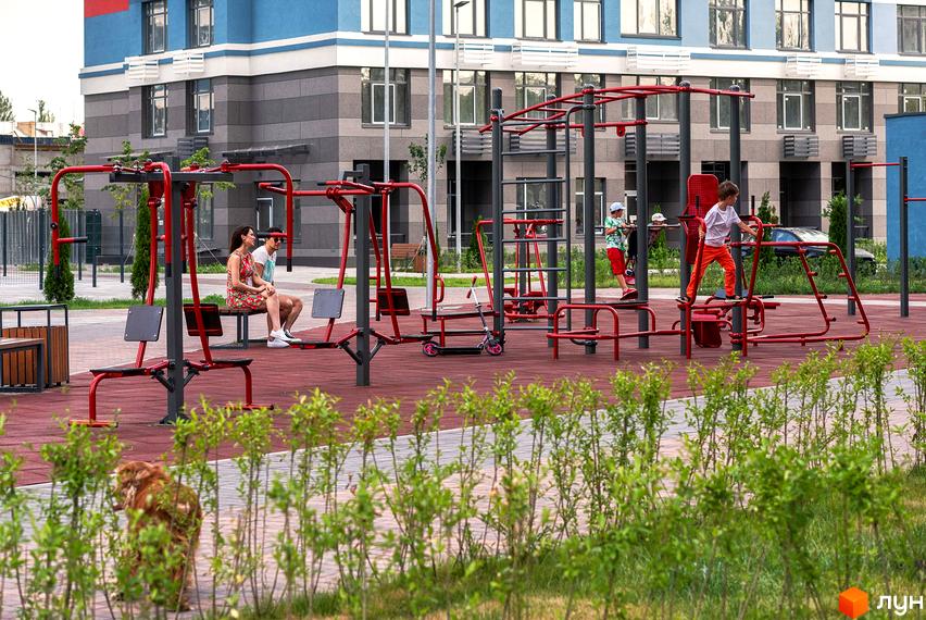 Житловий комплекс Галактика: дитячий майданчик з тренажерами та відпочиваючі біля багатоповерхових будинків. Зелена зона з кущами на передньому плані.