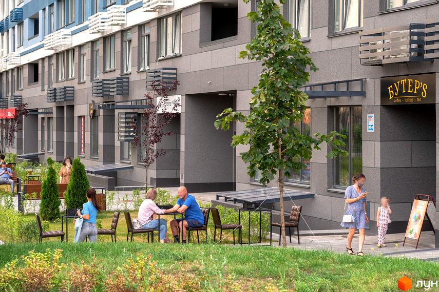 Житловий комплекс Галактика з сірим фасадом та зеленою прибудинковою територією. На вулиці розташовані столики та стільці для відпочинку.