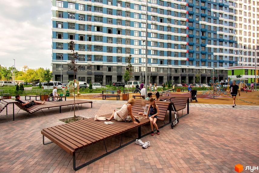 Житловий комплекс Галактика з синьо-білим фасадом та прибудинковою територією з дерев'яними лавами та дитячим майданчиком. Люди відпочивають на лавах біля будинку.