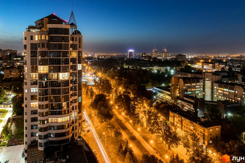 Житловий комплекс Славія на фоні нічного міста. Прибудинкова територія з освітленими алеями та зеленими насадженнями.