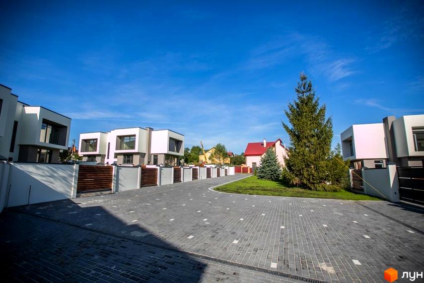 Котеджне містечко КМ Riviera Gold. Сучасні будинки з білими фасадами та темними вікнами, розташовані вздовж вимощеної сірою плиткою дороги з зеленими газонами та деревами.