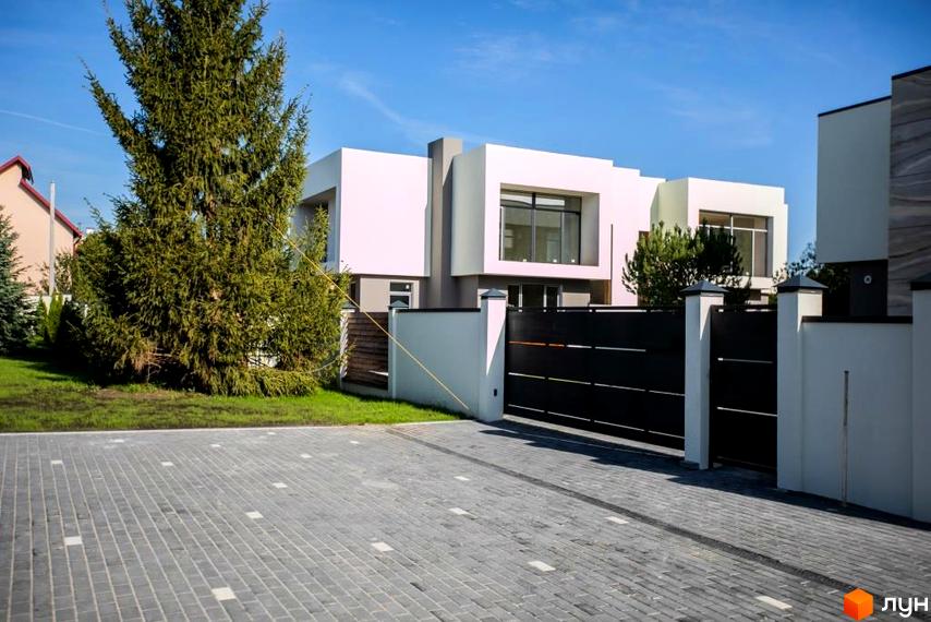 Котеджне містечко КМ Riviera Gold. Зовнішній вигляд будинку з білою штукатуркою та великими вікнами, огороджений чорним парканом. Прибудинкова територія з бруківкою та зеленими насадженнями.