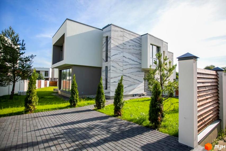 Котеджне містечко КМ Riviera Gold: двоповерховий будинок з сірою та білою обробкою, великими вікнами та дерев'яним парканом. Прибудинкова територія з газоном та бруківкою.