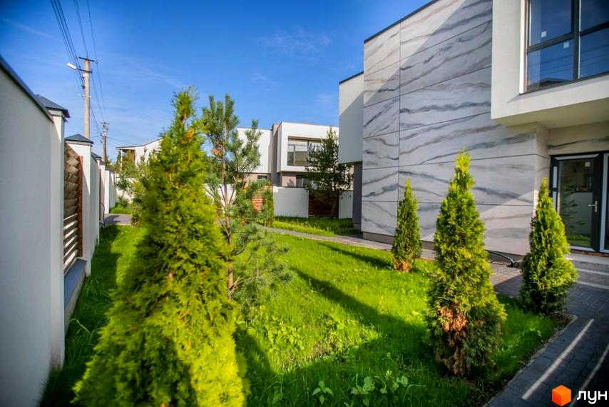 Зовнішній вигляд будинків КМ Riviera Gold з фасадами під мармур та зеленою прибудинковою територією з туями. Сучасний житловий комплекс з доглянутим ландшафтом.