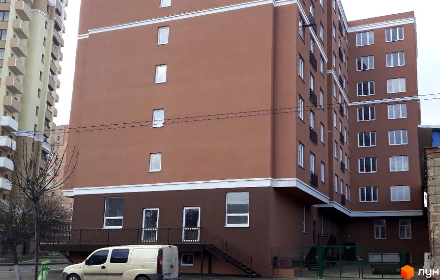 Зовнішній вигляд новобудови ЖК Слов'янський з коричневим фасадом та численними вікнами. Поруч видно прибудинкову територію з деревами та автомобілем.