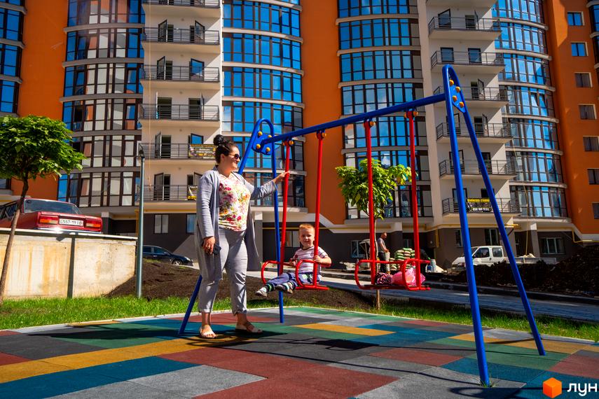 Зображення будинку Житловий комплекс Містечко Соборне з дитячим майданчиком. На передньому плані жінка з дитиною на гойдалці.
