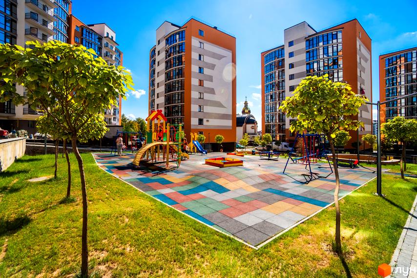 Зображення житлового комплексу Містечко Соборне з дитячим майданчиком. На фото висотні будинки та кольорове покриття ігрової зони.