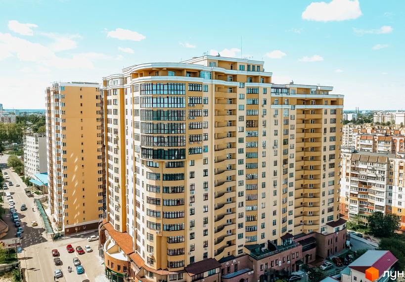 Житловий комплекс Авіа Квартал - висотна жовта новобудова з великою кількістю вікон та балконів. Навколо будинку розташована дорога з автомобілями та зелені насадження.