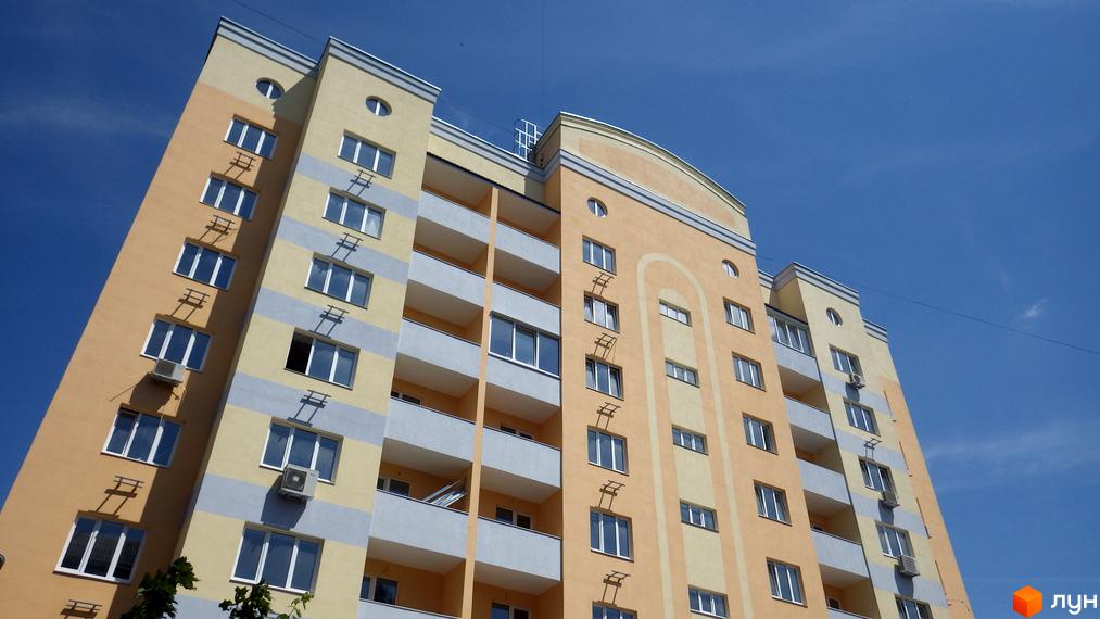 Зовнішній вигляд багатоповерхового будинку Клубний будинок Gelios з жовто-сірими фасадами та балконами на фоні блакитного неба. Новобудова з багатьма вікнами.