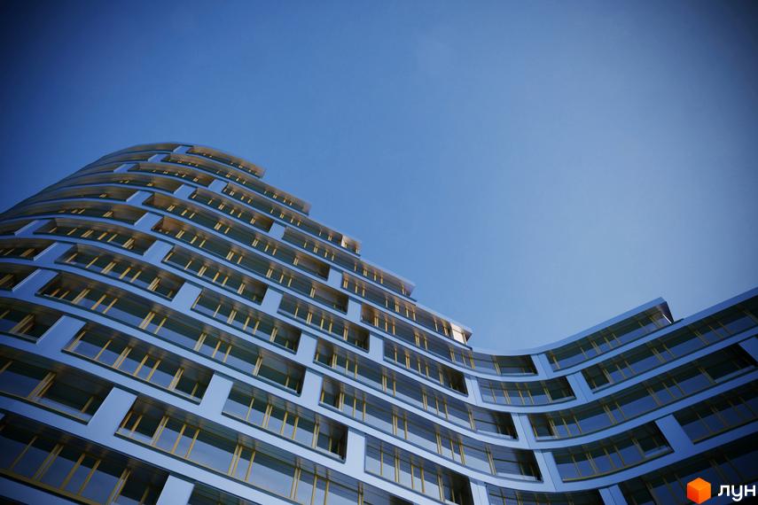 Житловий комплекс Alpina з вигнутим фасадом та численними вікнами на тлі блакитного неба. Зовнішній вигляд новобудови.