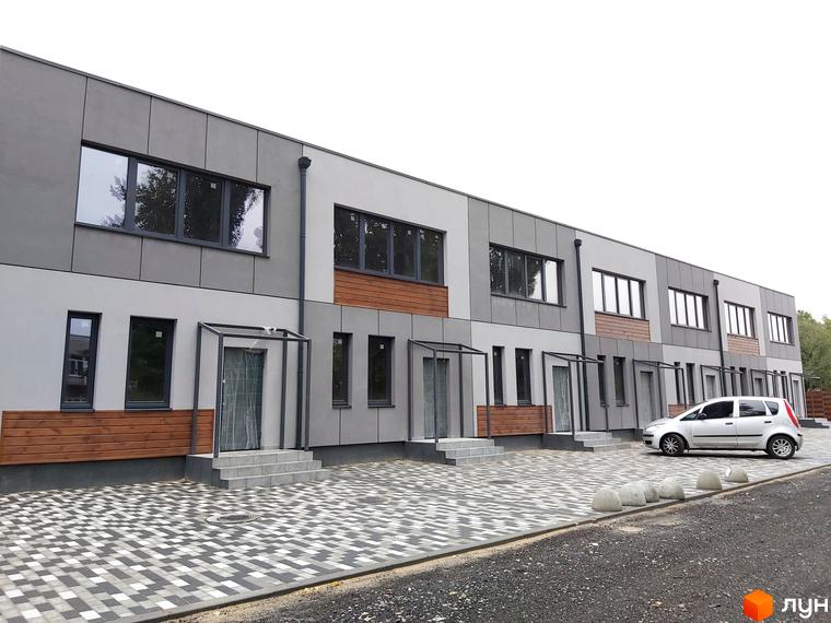 Зовнішній вигляд новобудови на вул. Сергія Колоса, 169, з сірими фасадами та дерев'яними вставками. Прибудинкова територія з бруківкою та автомобілем.