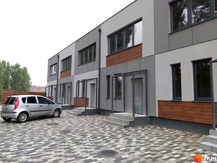 Зовнішній вигляд новобудови на вул. Сергія Колоса, 169, з сірими та білими фасадами та дерев'яними вставками. Прибудинкова територія вимощена плиткою, поруч припаркований сріблястий автомобіль.