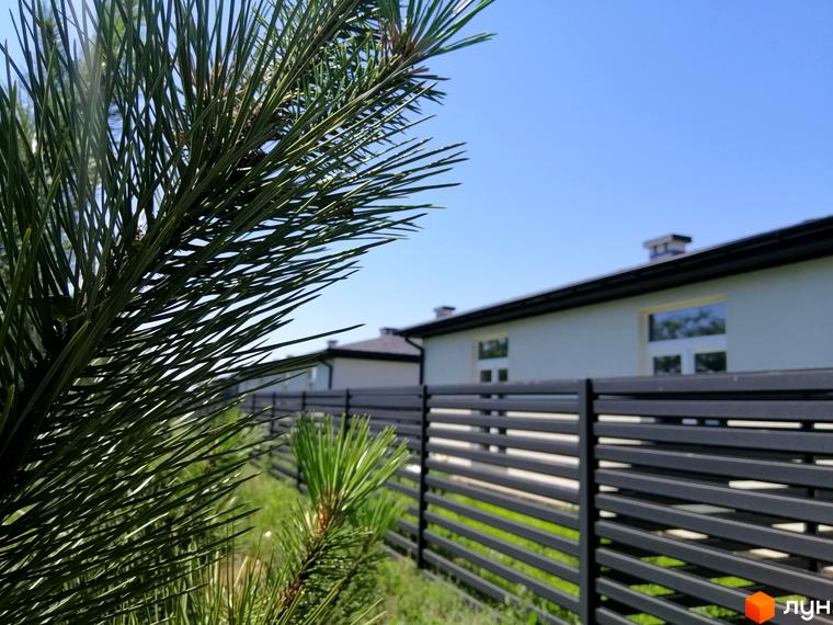 Фасад будинку Котеджного містечка Світлий Совіньон зі світлими стінами та темним дахом. На передньому плані темний горизонтальний паркан та зелена територія.