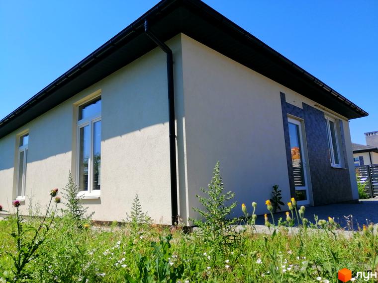 Містечко котеджів Світлий Совіньон: світлий одноповерховий будинок з темним дахом та білими вікнами під ясним небом. Перед ним зелена трава з квітами.