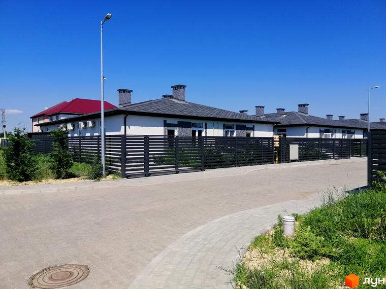 Будинки котеджного містечка Світлий Совіньон з білими фасадами та темними дахами, загороджені чорним парканом. На передньому плані бруківка та зелена трава.