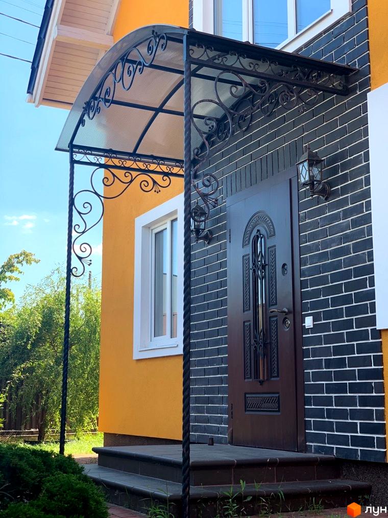 Котеджне містечко Osocor Scandi House: фасад будинку з помаранчевою штукатуркою, темною цеглою, кованим навісом над входом та зеленими насадженнями.