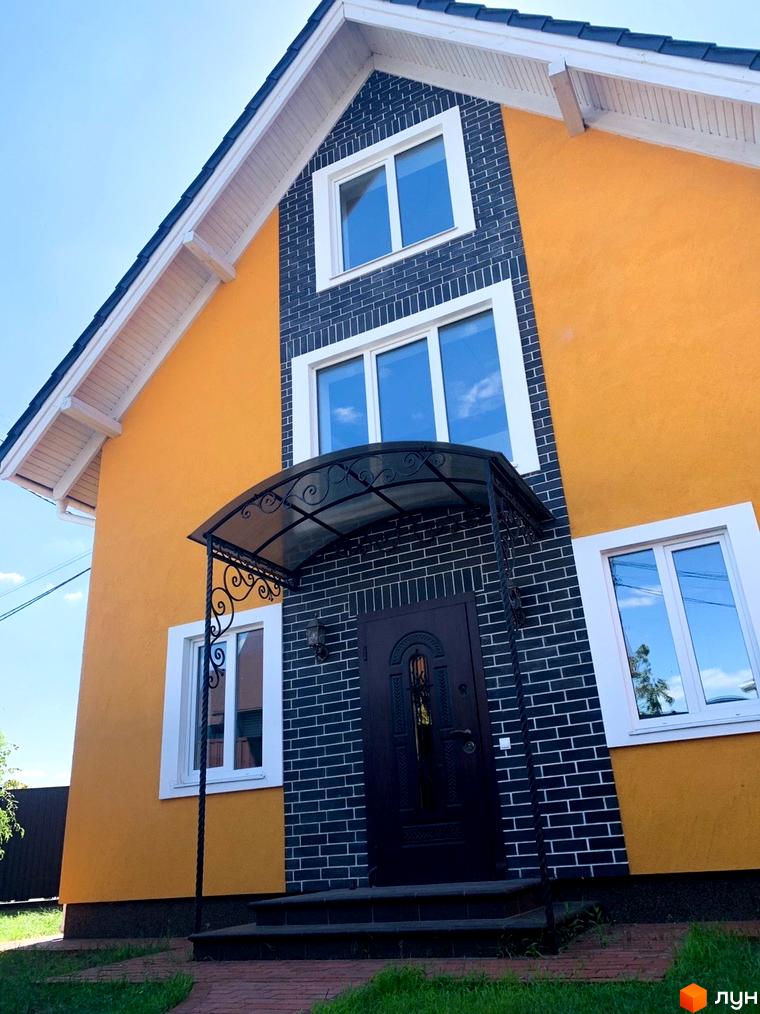 Фасад двоповерхового будинку з жовтими стінами та темно-сірою цегляною кладкою. Перед входом сходи та кований навіс.