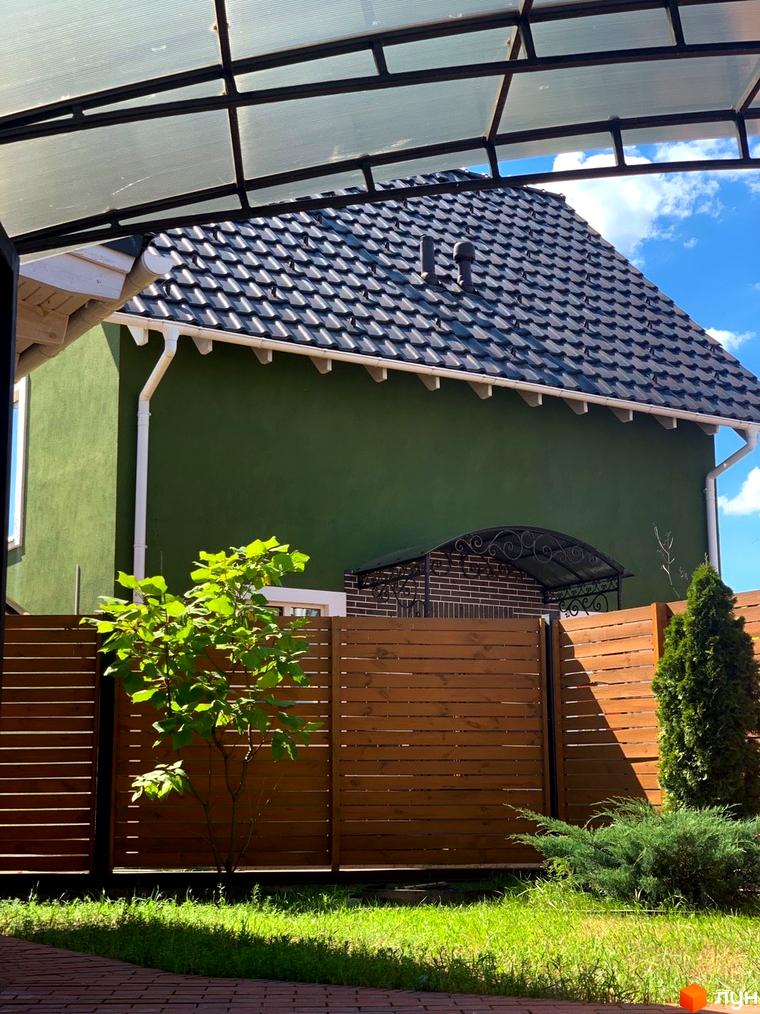 Вигляд будинку в Котеджному містечку Osocor Scandi House з чорною черепицею та зеленою стіною. Навколо зелена трава та дерев'яний паркан.