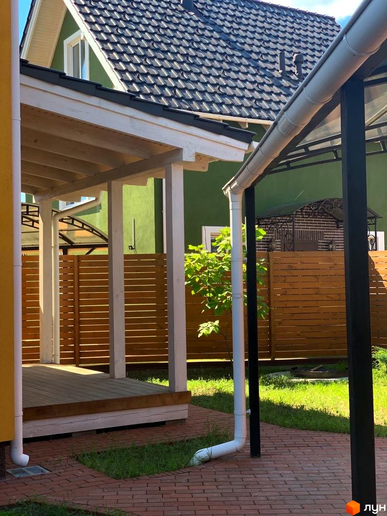 Зелений фасад будинку Котеджне містечко Osocor Scandi House з темно-сірою покрівлею та білим ґанком. Прибудинкова територія з бруківкою та зеленим газоном.