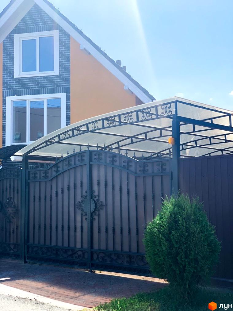 Будинок Котеджного містечка Osocor Scandi House з помаранчевим фасадом та сірою цегляною обробкою, перед яким розташовані металеві ворота з прозорим навісом та зелений кущ.