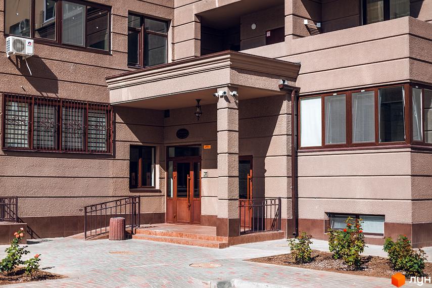 Зовнішній вигляд житлового комплексу Одеські традиції. Фасад будинку з вікнами, вхідна група з колонами та сходинками, прибудинкова територія з тротуарною плиткою.