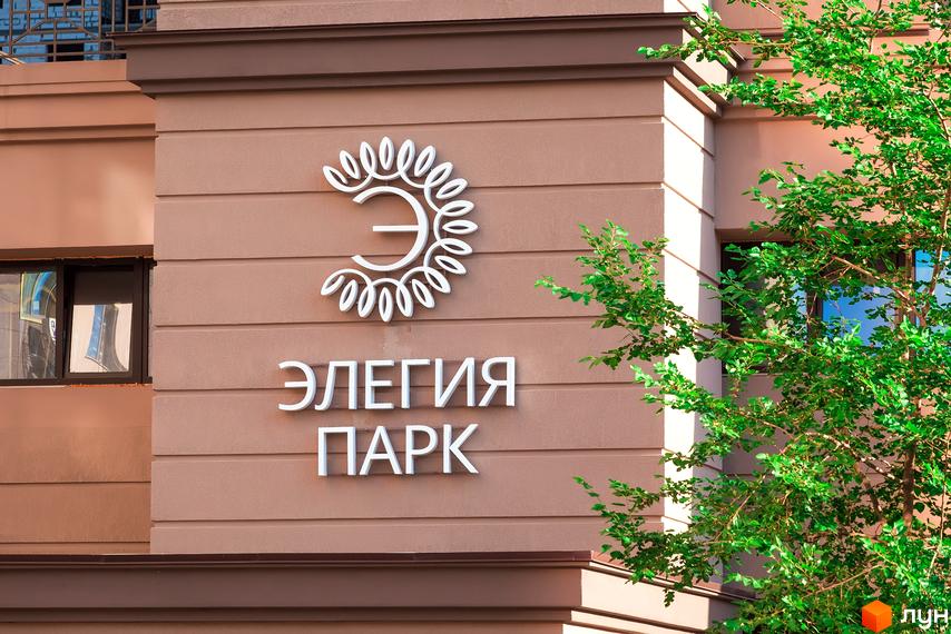 Фасад житлового комплексу Елегія Парк з білим логотипом та зеленою рослинністю. Зовнішній вигляд новобудови з чіткими лініями та текстурними елементами.