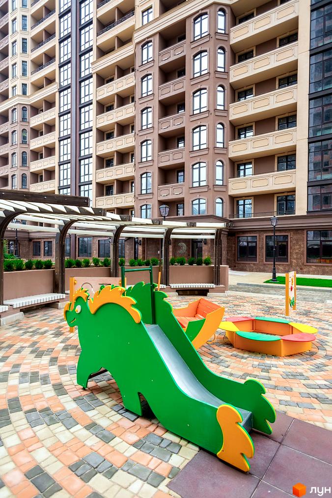 Елегія Парк: фасади будинків з балконами та дитячий майданчик з гіркою та пісочницею.