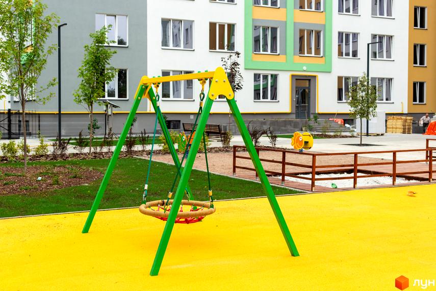 Житловий комплекс 3V Volkova. Дитячий майданчик з гойдалкою та яскравим покриттям на тлі багатоповерхових будинків.