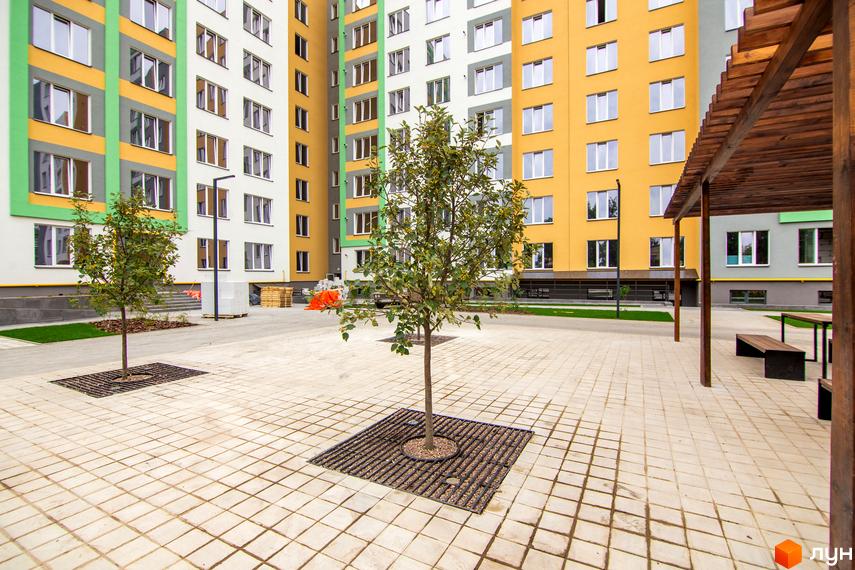 Житловий комплекс 3V Volkova: новобудова з яскравими фасадами жовтого, білого та зеленого кольорів. На прибудинковій території розташовані дерева, лавки та плитка.
