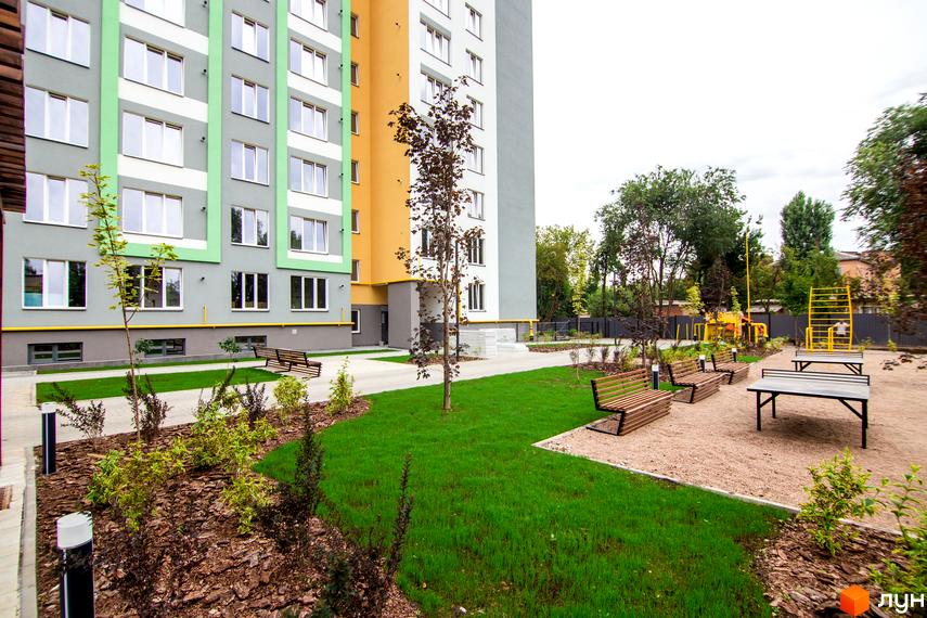Житловий комплекс 3V Volkova. Фасад будинку з сірими та зеленими елементами, прибудинкова територія з лавками та дитячим майданчиком.