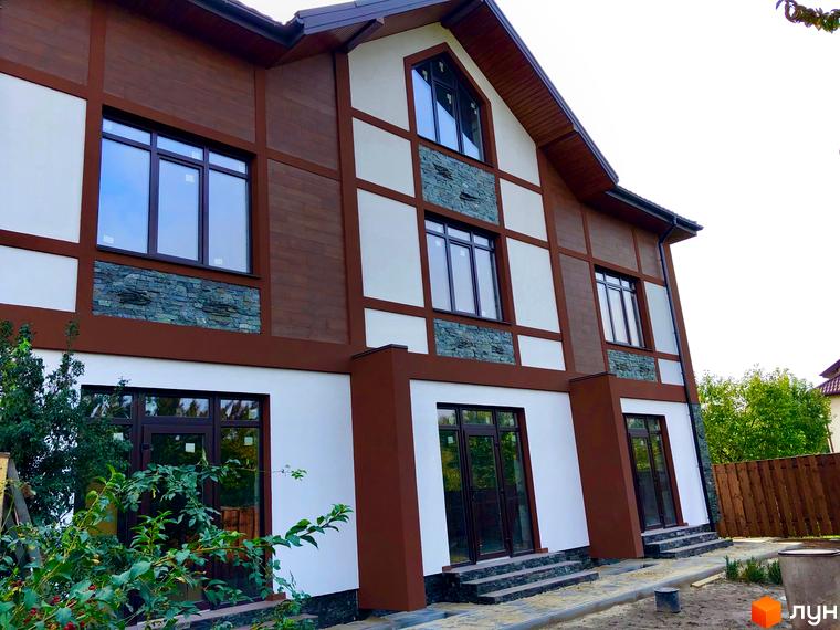 Зовнішній вигляд таунхаусів Київ Шале з оздобленням фасаду під дерево та камінь. На передньому плані зелені насадження.