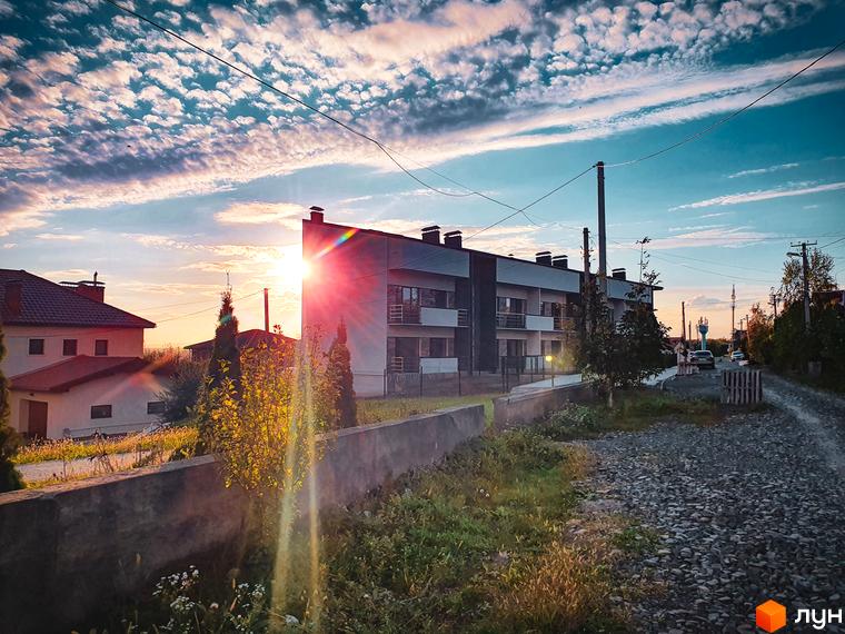 Житловий комплекс Клубний з фасадами сірого кольору та червоними дахами на фоні неба з хмарами. Територія навколо комплексу вкрита зеленню та гравієм.