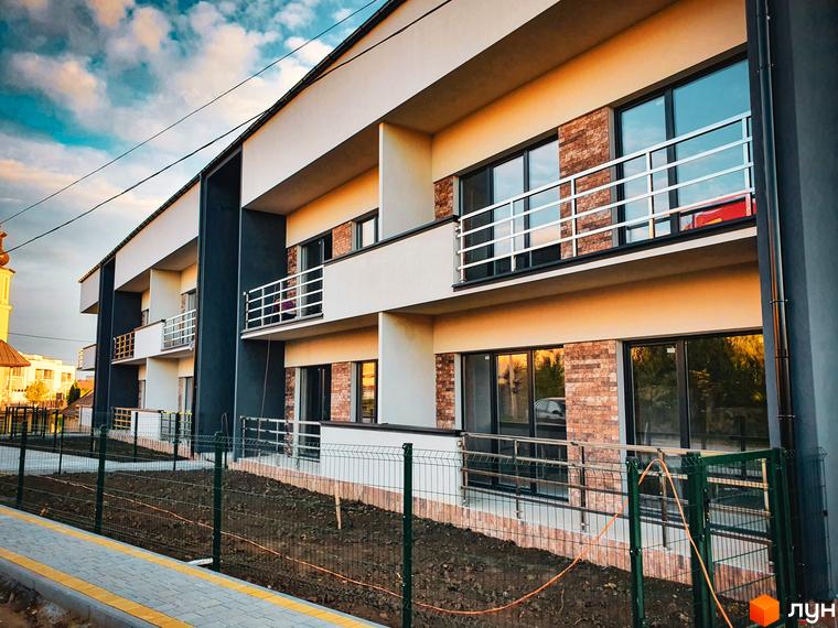 Зовнішній вигляд ЖК Клубний з балконами та цегляним оздобленням фасаду. Територія навколо будинку огороджена парканом, видно тротуар та зелені насадження.